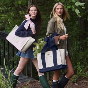 Two women standing outdoors in a wooded setting, each carrying a large tote bag with a wide ribbon handle. The tote bags are cream with navy blue panels and handles. #color_ballet pink bow