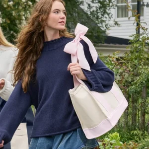 A woman outdoors wearing a navy knit sweater and holding a cream tote bag with a large light pink ribbon bow at the top. #color_ballet pink bow