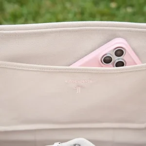 Close-up of a beige canvas shoulder bag with a pink smartphone tucked into the side pocket. #color_ballet pink bow
