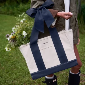 A person outdoors carrying a large off-white tote bag with bold navy blue side panels and handles, tied with a matching navy bow. The tote has a front pocket with the visible brand name 'CASE-MATE' and is filled with wildflowers. #color_oxford navy bow