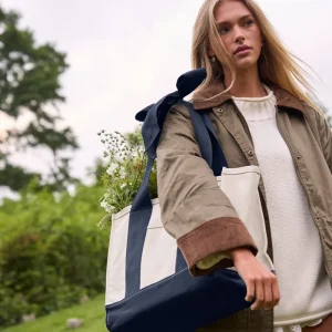 A woman outdoors carrying a large cream and navy tote bag over her shoulder, with a white sweater and olive jacket visible. The background shows blurred greenery and sky. #color_oxford navy bow