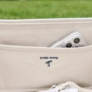 Close-up of a beige canvas laptop sleeve with the CASE-MATE logo embroidered in navy on the front, showing a silver smartphone tucked into a side pocket, with a blurred green grass background. #color_oxford navy bow
