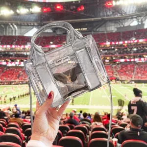 Clear handbag with 'CASE-MATE' branding held in front of a sports stadium. #color_silver chrome